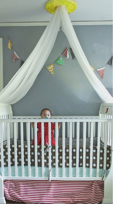 Beautiful baby girl standing in her crib located in a colorful shared nursery with gray walls and a yellow ceiling medallion